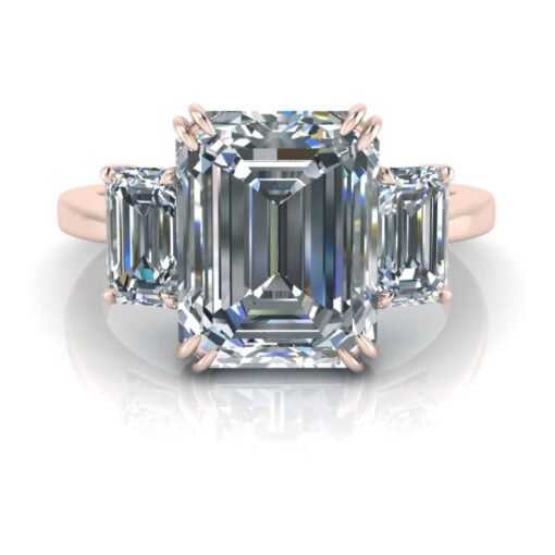 A rose gold three stone emerald cut engagement ring laying on flat reflective surface.